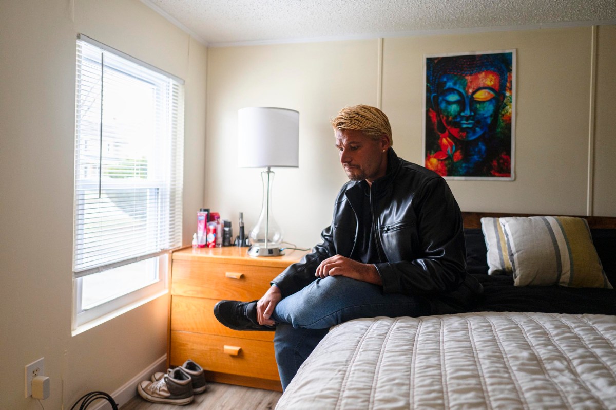 A person with blond hair in a black leather jacket sits on the edge of a bed in a bedroom, looking down, beside a window and a dresser with shoes on the floor.