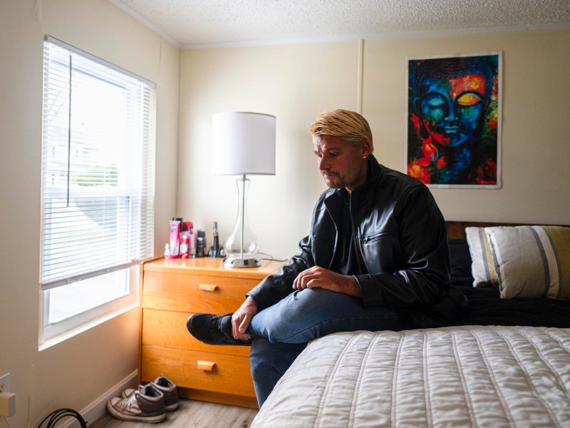 A person with blond hair in a black leather jacket sits on the edge of a bed in a bedroom, looking down, beside a window and a dresser with shoes on the floor.
