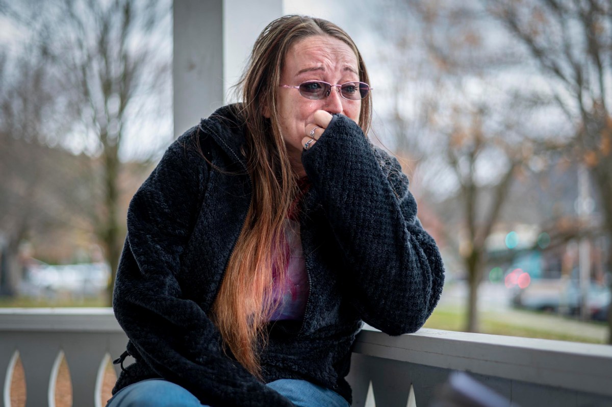 A woman with long hair and glasses sits on a porch, visibly upset, covering her mouth with her hand and appearing to cry.