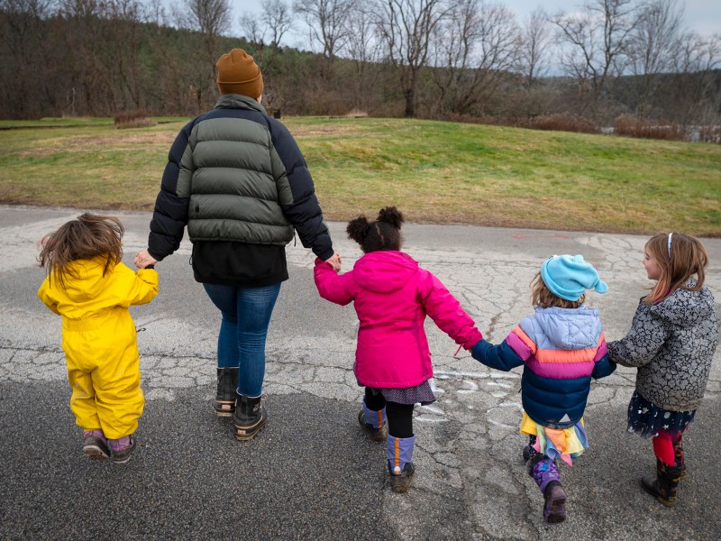 An adult and four young children, dressed in colorful winter clothes, walk hand-in-hand on a paved path with grass and trees in the background.
