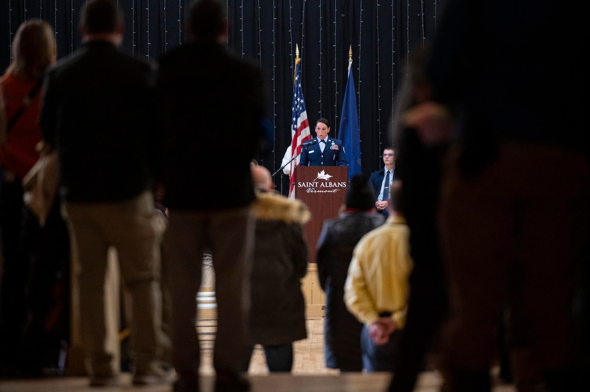 A person in military uniform speaks at a podium labeled "Saint Albans" with two flags behind them, while an audience listens in the foreground.