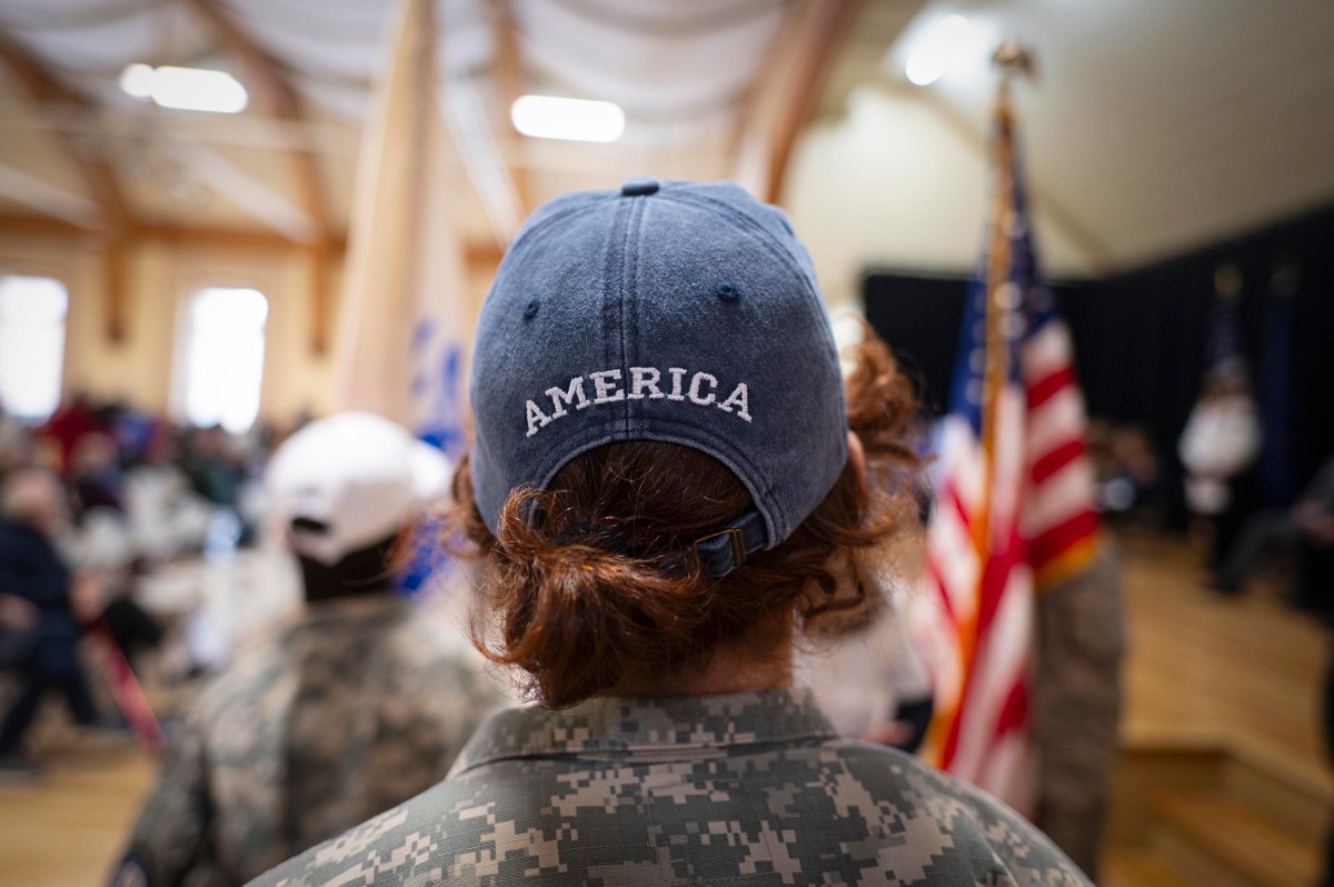 Person in military uniform with long hair, seen from behind, wearing a blue cap labeled "AMERICA," standing indoors near an American flag.