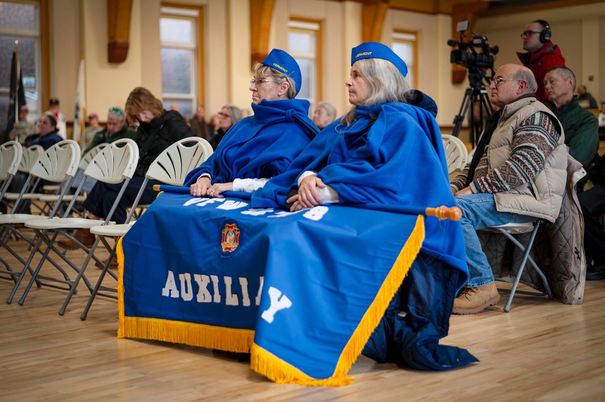 Two women in blue uniforms and capes labeled "AUXILIARY" sit in folding chairs during an indoor event, with other attendees and a cameraman in the background.