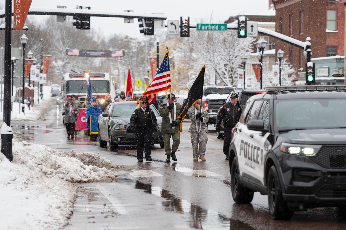 A group of people, including uniformed individuals, carry flags and march down a snowy street, escorted by a police vehicle.