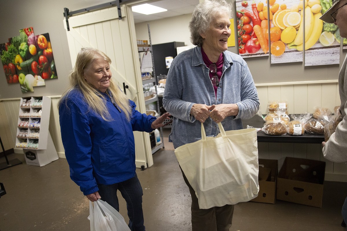 Two women stand indoors holding shopping bags, one smiling and the other looking off to the side. Shelves, baked goods, and colorful fruit-themed art are visible in the background.