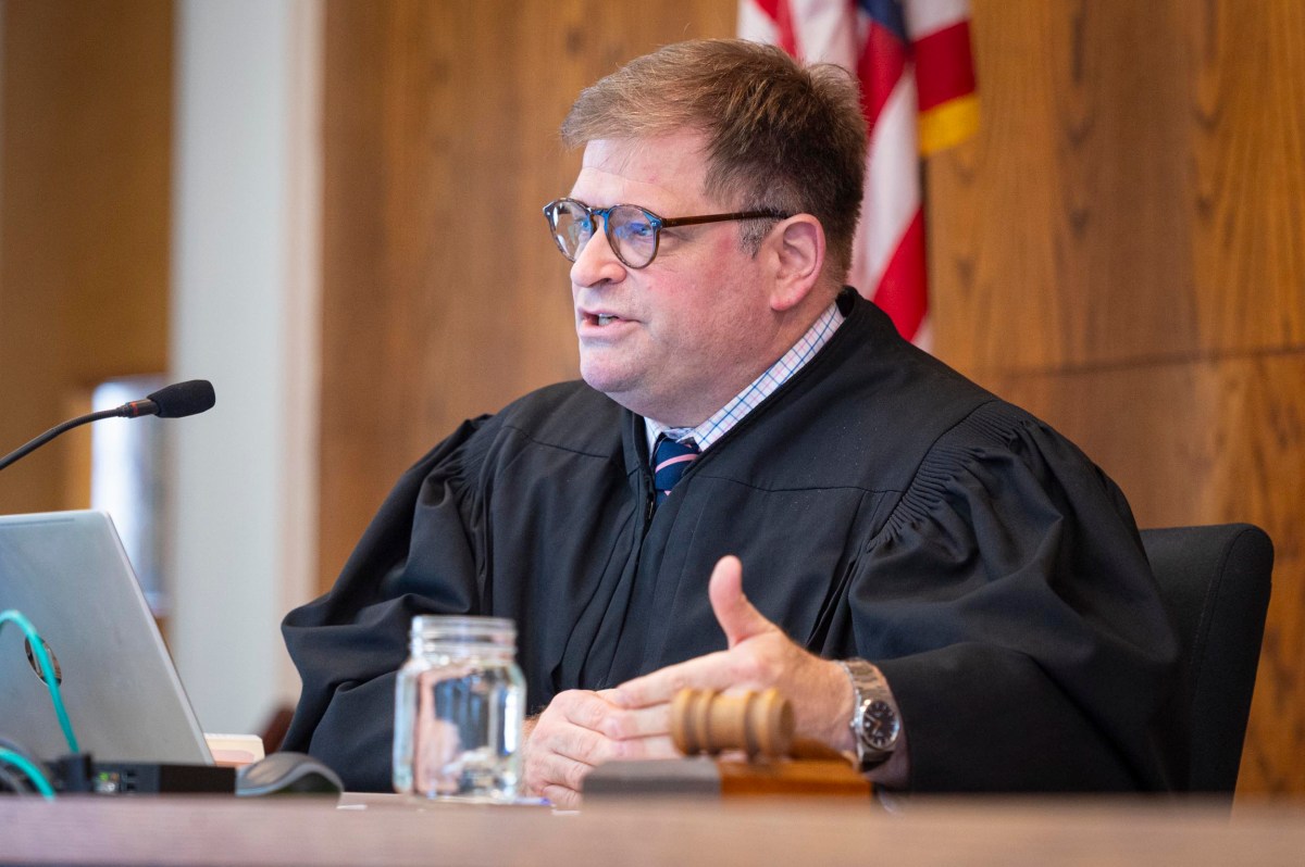 A judge wearing black robes sits at a bench with a laptop, microphone, gavel, and water jar, speaking in a courtroom. An American flag is visible in the background.