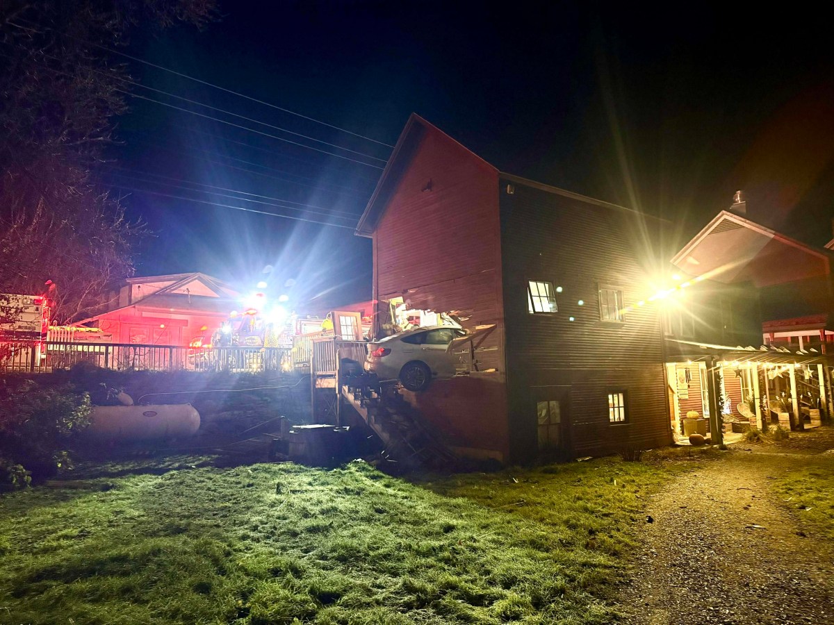 A car is lodged on the wooden stairs outside a house at night, with emergency vehicles and bright lights in the background.