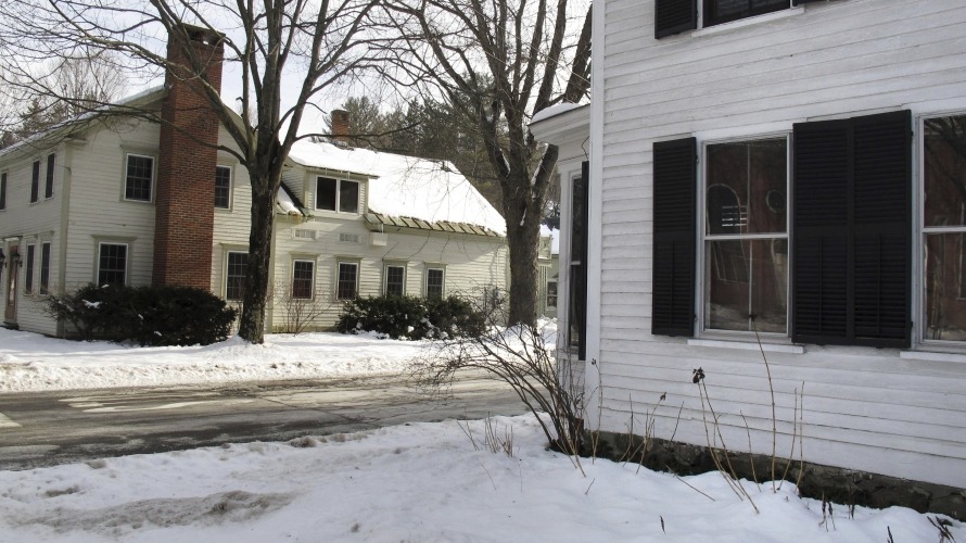 Two white wooden houses with black shutters are shown on a snowy street in winter, with bare trees and snow covering the ground.