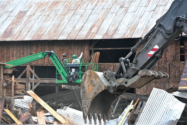 Two workers in a green lift operate next to a large excavator claw amid demolished wood and metal debris in front of a wooden building.