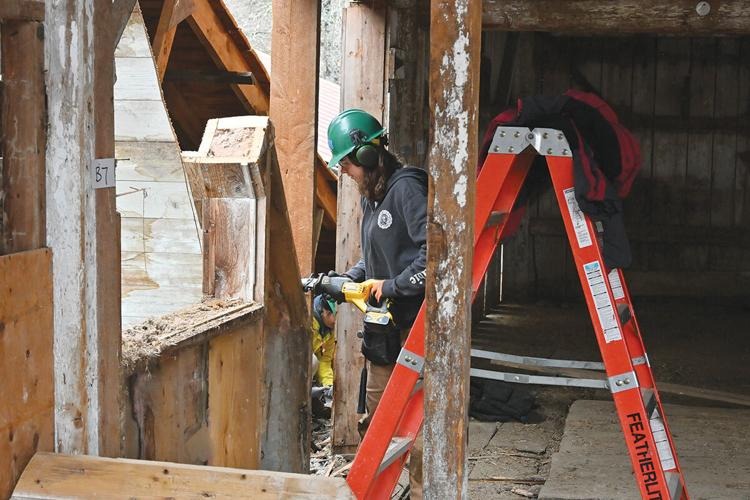 A person in a hard hat and safety gear uses a power tool to work on wooden beams inside a partially constructed or renovated building beside an orange ladder.