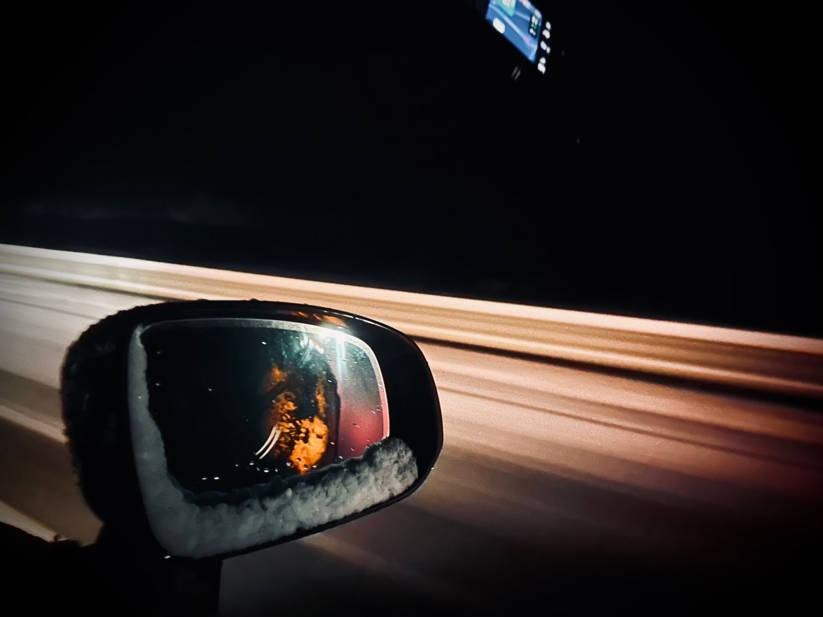 A car’s side mirror reflects an illuminated face and hands, with blurred lights from a road at night visible in the background.