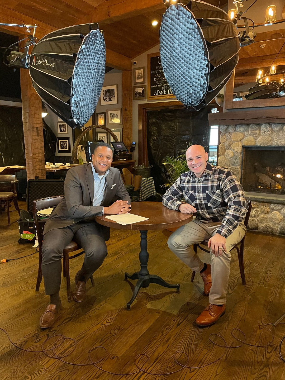 Two men sit at a round table in a rustic indoor setting with professional lighting equipment overhead, preparing for what appears to be an interview or discussion.