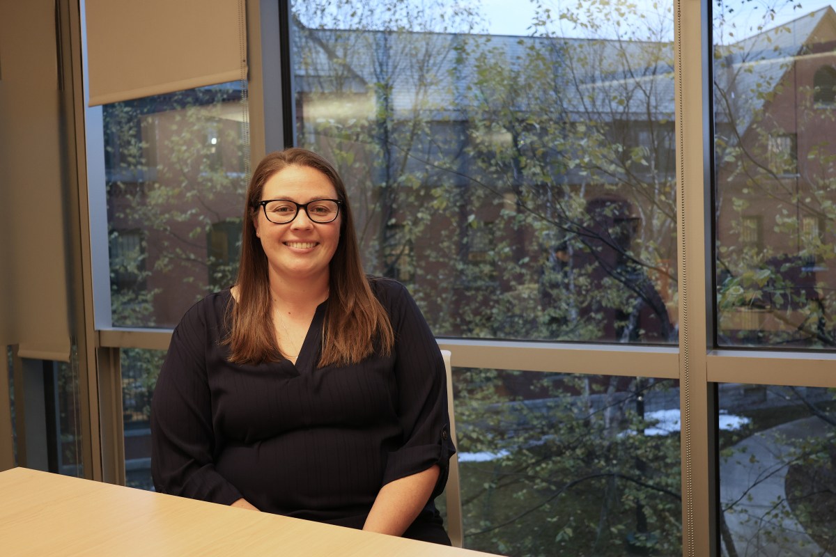 A woman with long brown hair and glasses sits at a desk in an office with large windows overlooking trees and a building outside.
