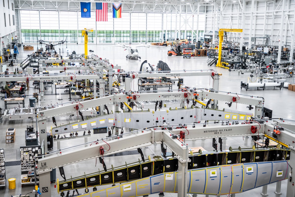 Wide view of a modern aerospace manufacturing facility with large aircraft fuselage sections, machinery, and assembly stations under bright industrial lighting.