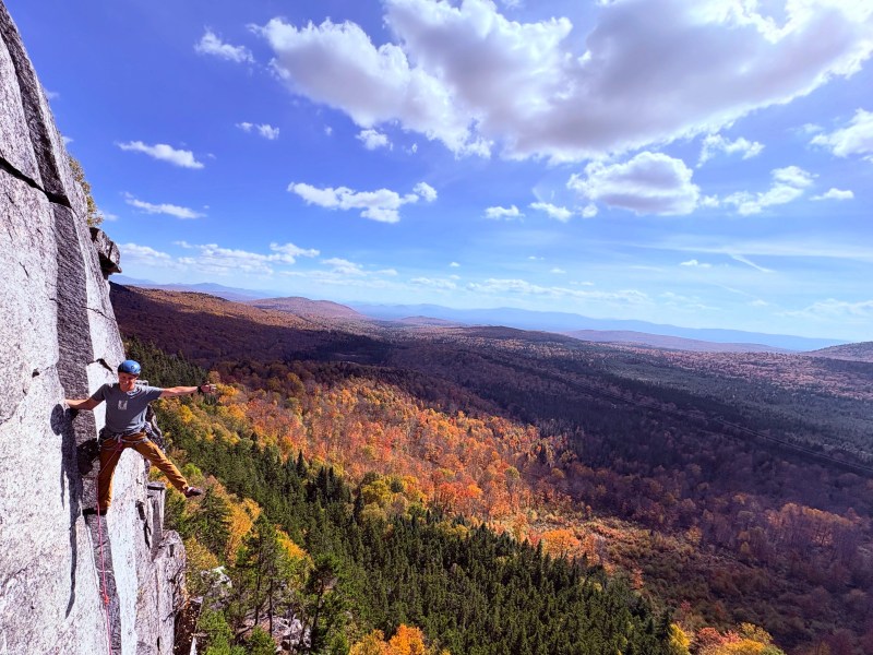 A person rock climbing on a cliff with a scenic view of a forested valley and distant mountains under a partly cloudy sky.