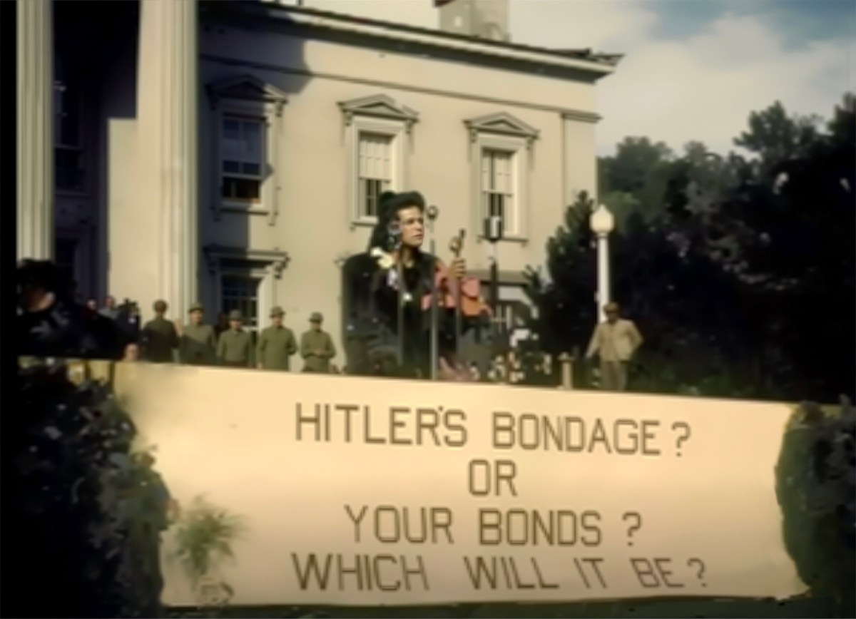 A speaker addresses a crowd from a platform with a large banner reading, "HITLER'S BONDAGE? OR YOUR BONDS? WHICH WILL IT BE?" in front of a building.