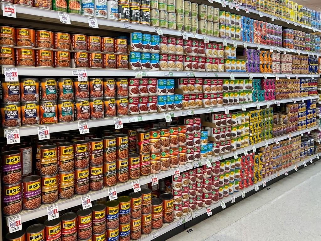 Shelves in a grocery store stocked with various brands and types of canned beans, soups, and vegetables in colorful packaging.