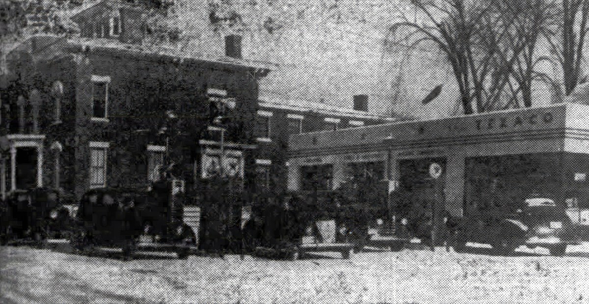 Black and white photo of several cars parked outside a brick building and a Texaco gas station on a snowy street.