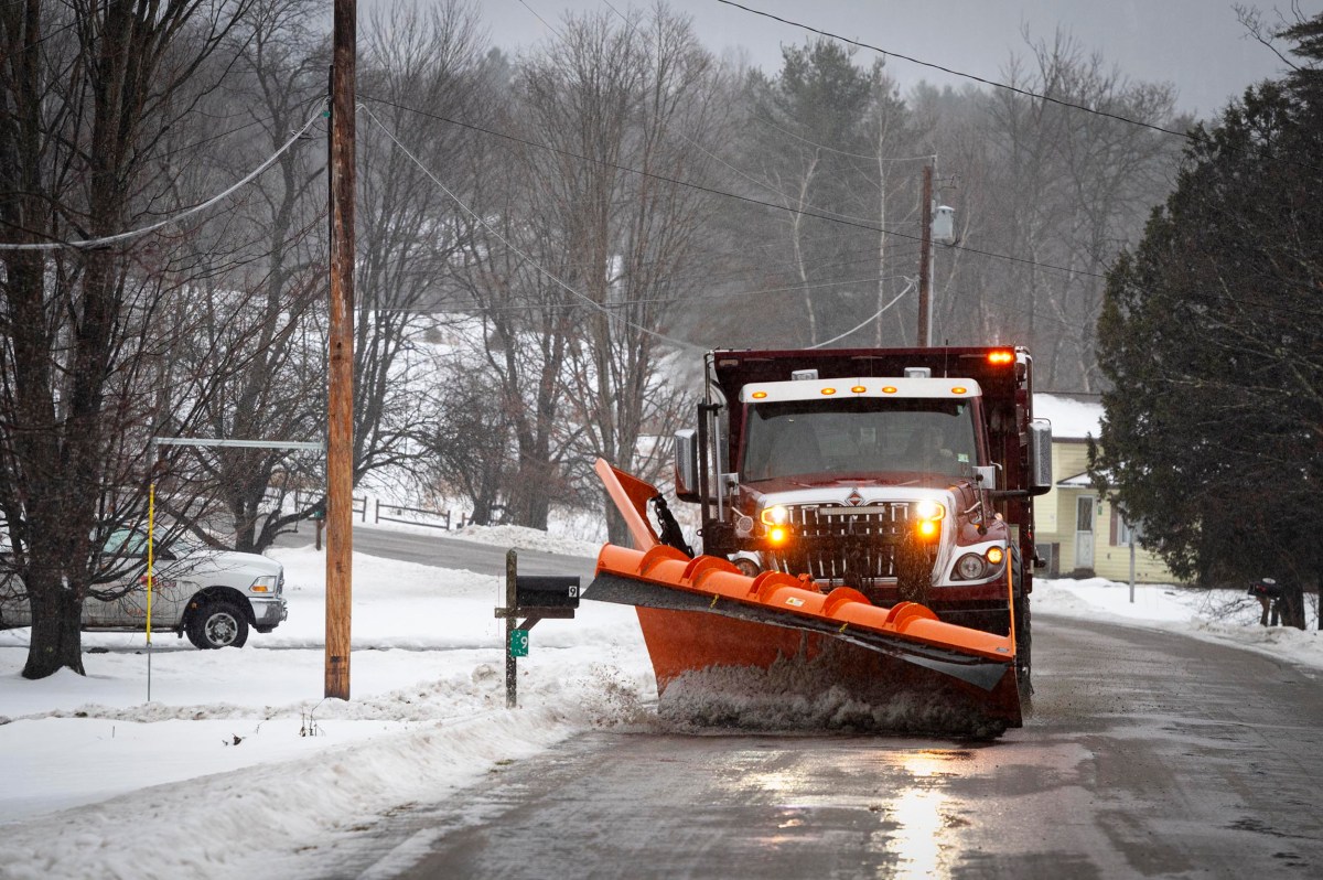 A snowplow clears snow from a residential road on a snowy day, with a pickup truck parked nearby and snow-covered trees in the background.