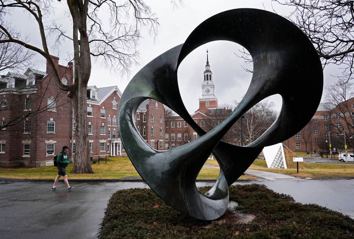 A person walks past a large abstract metal sculpture on a college campus, with brick buildings and a clock tower in the background on a cloudy day.