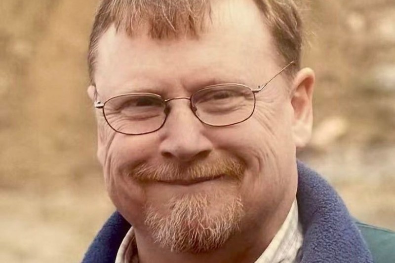 A man with glasses, short light brown hair, and a goatee smiles at the camera, wearing a plaid shirt and a blue fleece jacket.