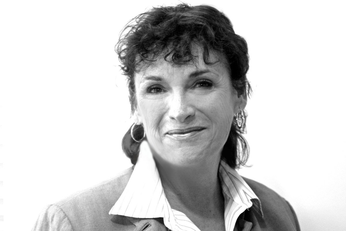 A woman with short, curly dark hair, wearing a collared shirt and blazer, smiles slightly at the camera against a plain white background.