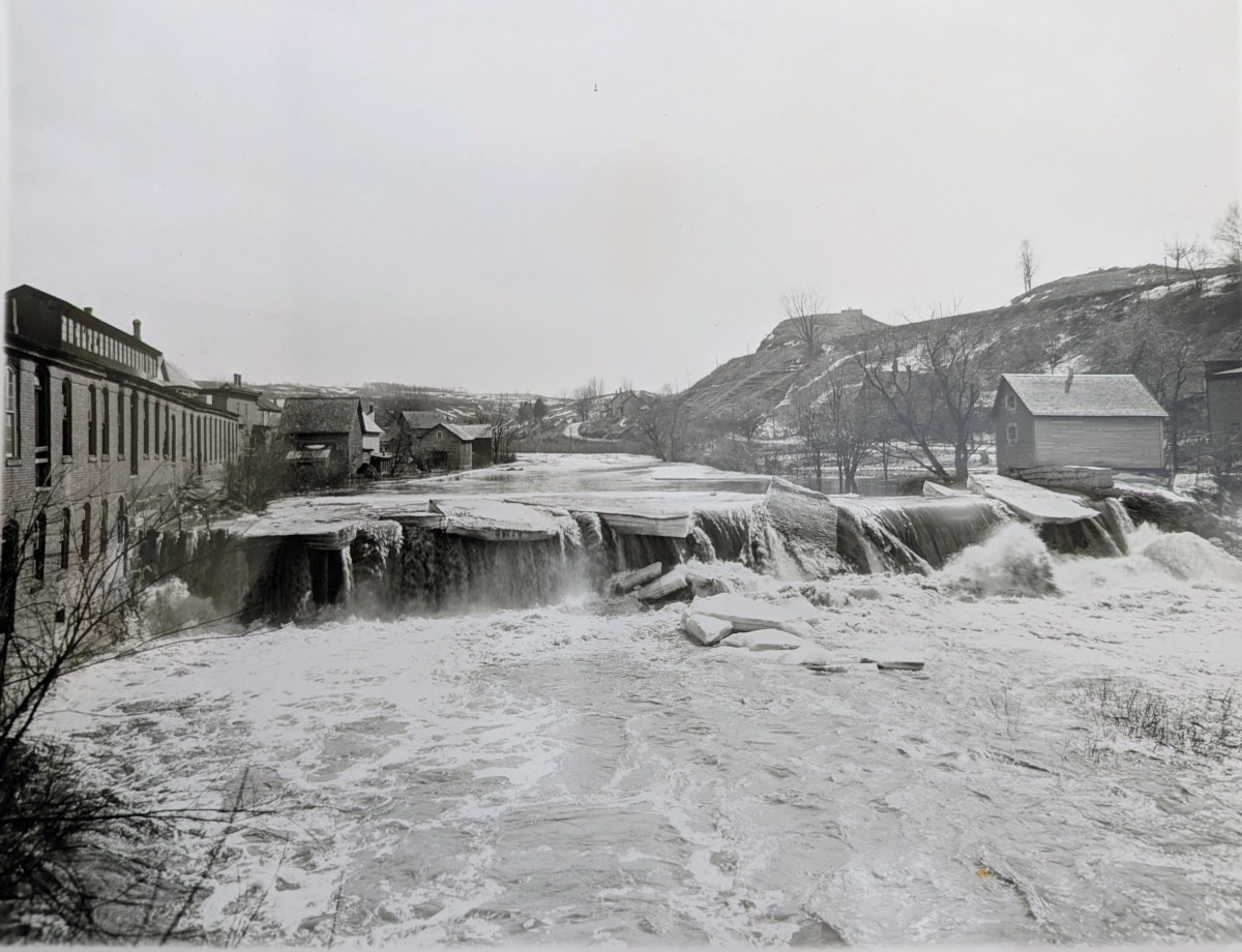 A river with a wide waterfall flows between buildings and bare trees, with ice and snow visible on the water and surrounding landscape.