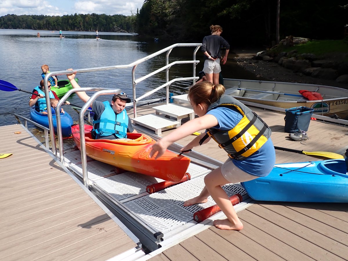 A girl in a life jacket helps launch a kayak with a man inside from a dock using a ramp system at a lakeside. Other people and kayaks are visible nearby.