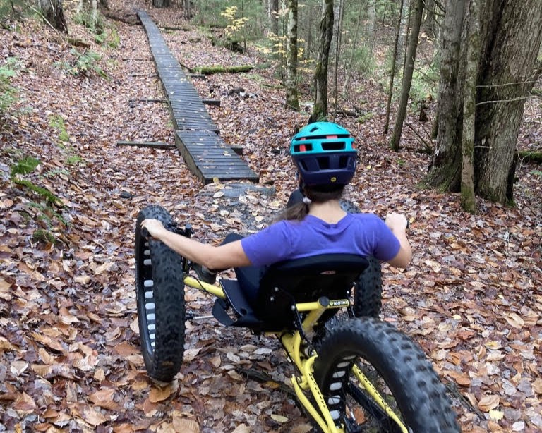 A person wearing a helmet rides a three-wheeled off-road adaptive bike on a leaf-covered forest trail toward a wooden boardwalk.