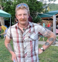 A man with a mustache stands outdoors, wearing a plaid shirt and sunglasses on his head, with tattoos visible on both arms. Trees and people are in the background.