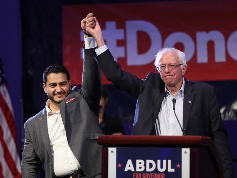 Two men stand on stage, one in a suit and the other in a jacket, holding hands raised in victory. An American flag and campaign signs are visible in the background.