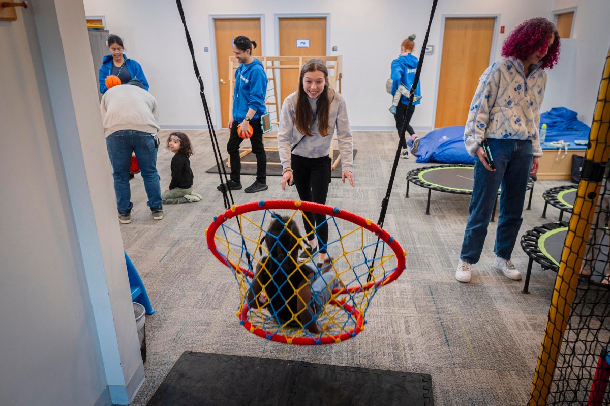 Children and adults interact in a playroom with trampolines, a swing, and toys; one child pushes another in a netted swing.