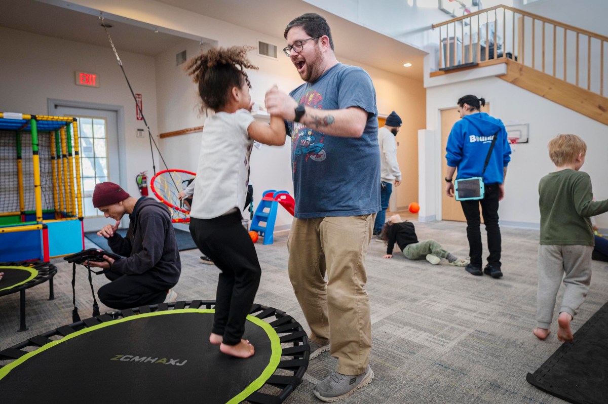 An adult and a child jump on a trampoline indoors while others play and interact in a brightly lit room with toys and play equipment.