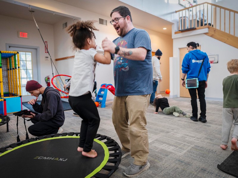 An adult and a child jump on a trampoline indoors while others play and interact in a brightly lit room with toys and play equipment.