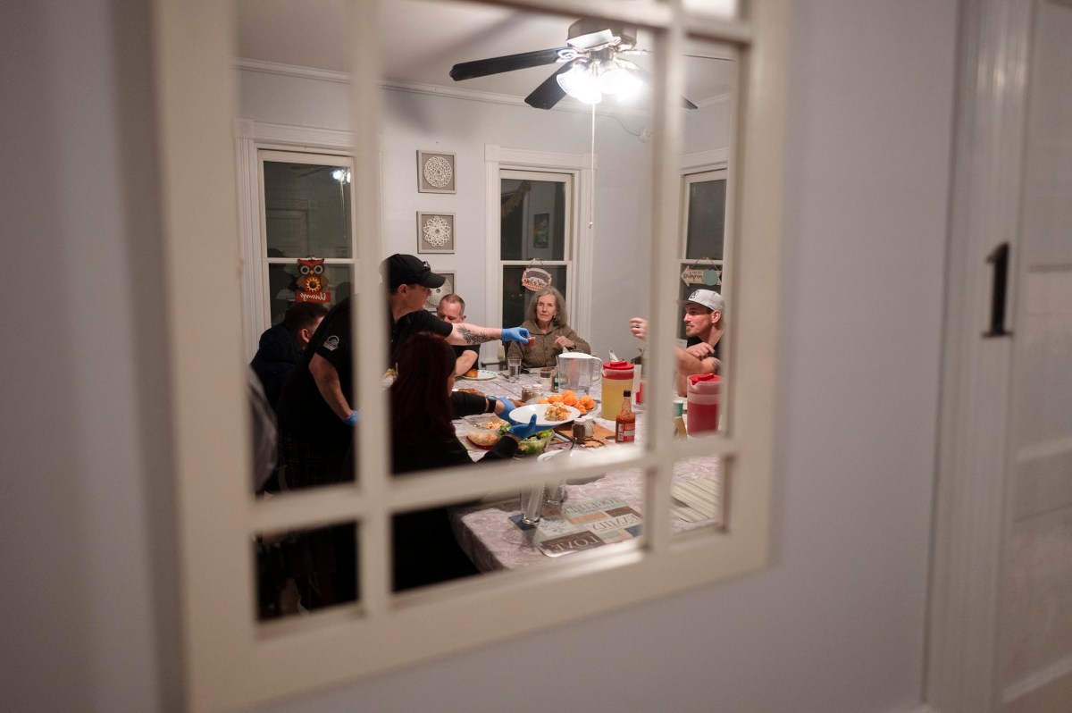A group of people are gathered around a dining table, sharing a meal and talking, as seen through a window from another room.