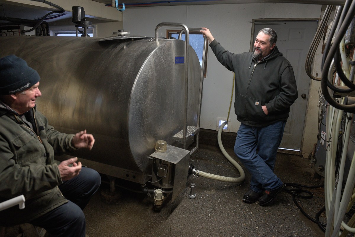 Two men talk next to a large stainless steel tank in an industrial indoor setting, with pipes and equipment visible around them.