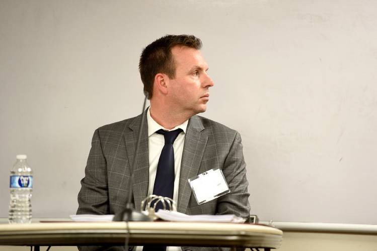 A man in a gray suit sits at a table with papers and a water bottle, looking to his left in front of a whiteboard.