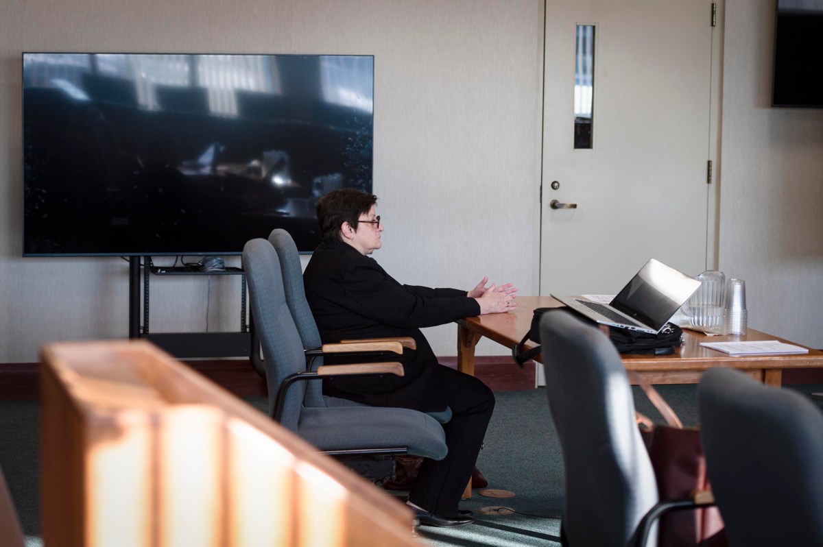 A person in a black suit sits alone at a table in a courtroom, facing a laptop, with empty chairs and a large screen in the background.