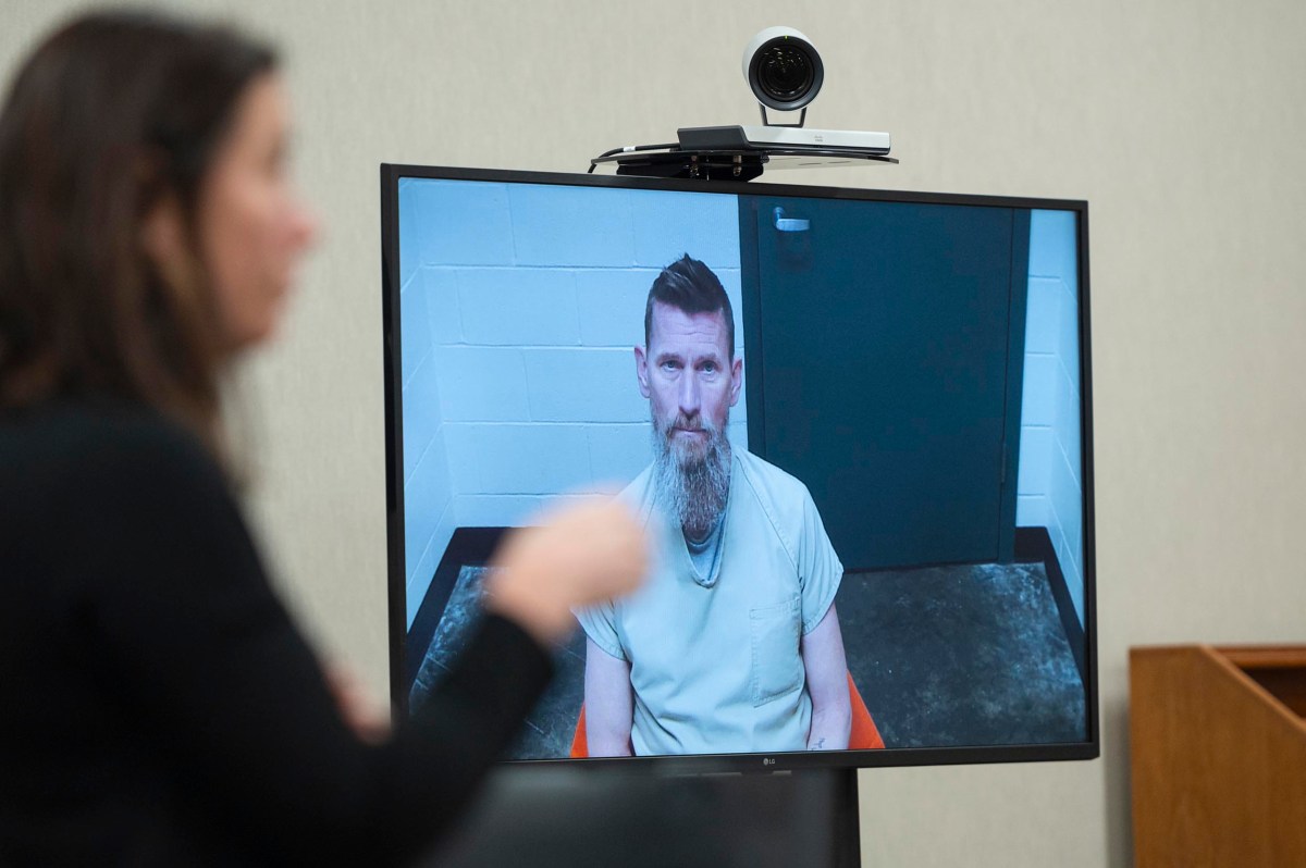 A man in a prison uniform appears on a video call screen during a remote court hearing, while a woman stands in the foreground.