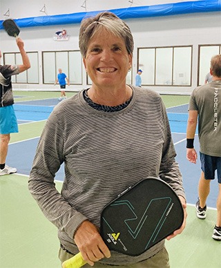 Smiling person holding a pickleball paddle stands on an indoor court with other players in the background.