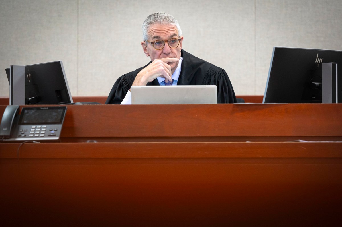 A judge wearing a black robe sits at a courtroom bench in front of computer monitors, appearing thoughtful.