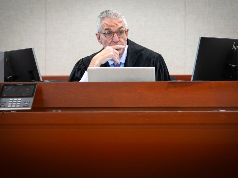 A judge wearing a black robe sits at a courtroom bench in front of computer monitors, appearing thoughtful.