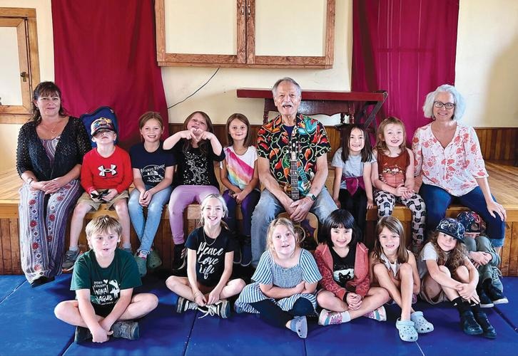 A group of children and three adults sit and pose for a group photo indoors, with wooden floors and red curtains in the background.
