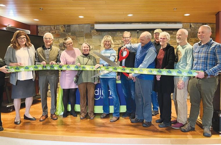 A group of people stands indoors, smiling and holding a large decorative ribbon while one person prepares to cut it with oversized scissors at a ribbon-cutting event.