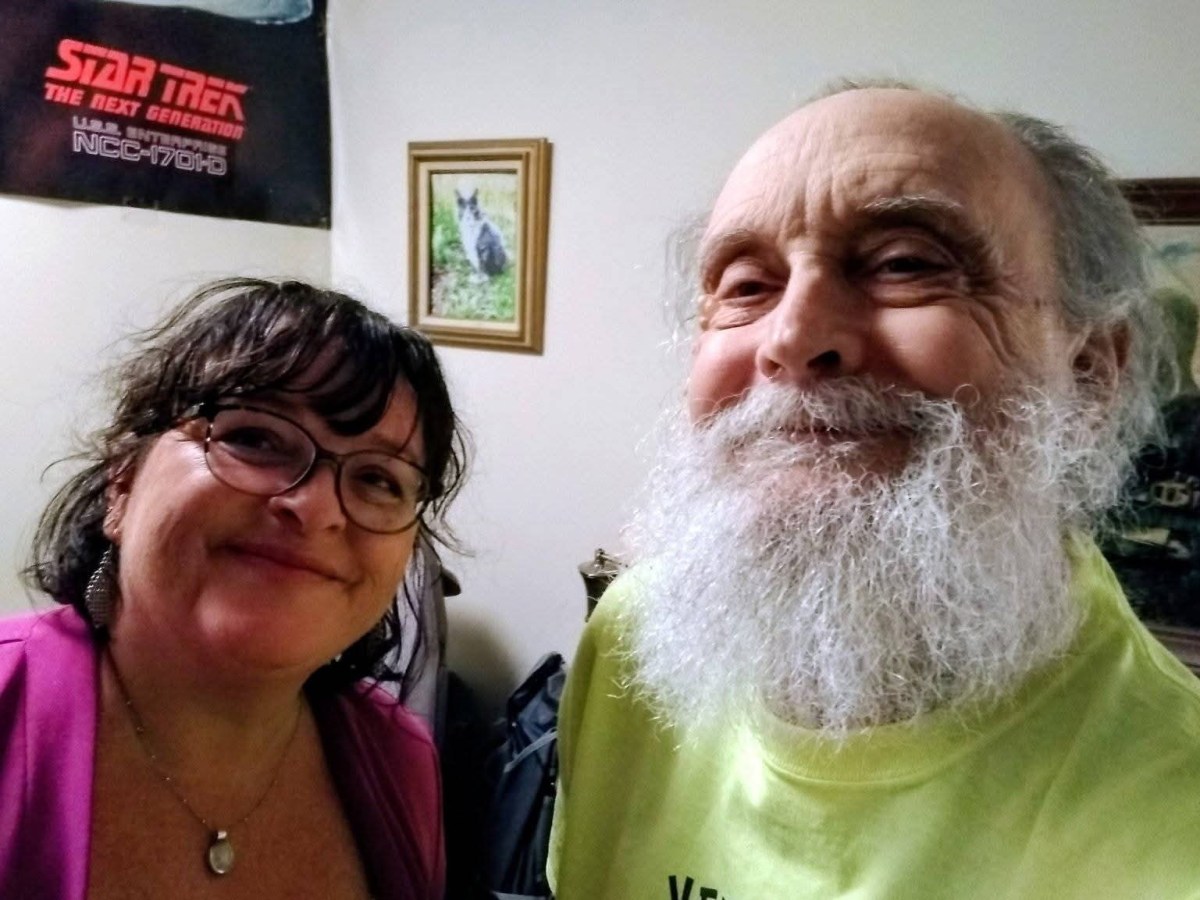 A woman with glasses and a man with a white beard smile indoors; a Star Trek: The Next Generation poster and framed artwork are visible in the background.