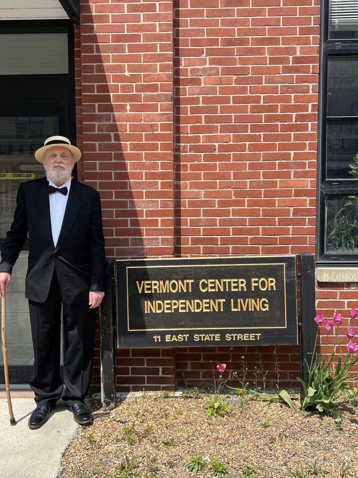 An older man in a suit and hat stands with a cane beside a sign reading "Vermont Center for Independent Living, 11 East State Street" in front of a brick building.