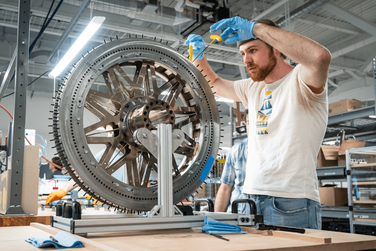 A man wearing blue gloves works on a large, mounted mechanical wheel using tools in a workshop setting.