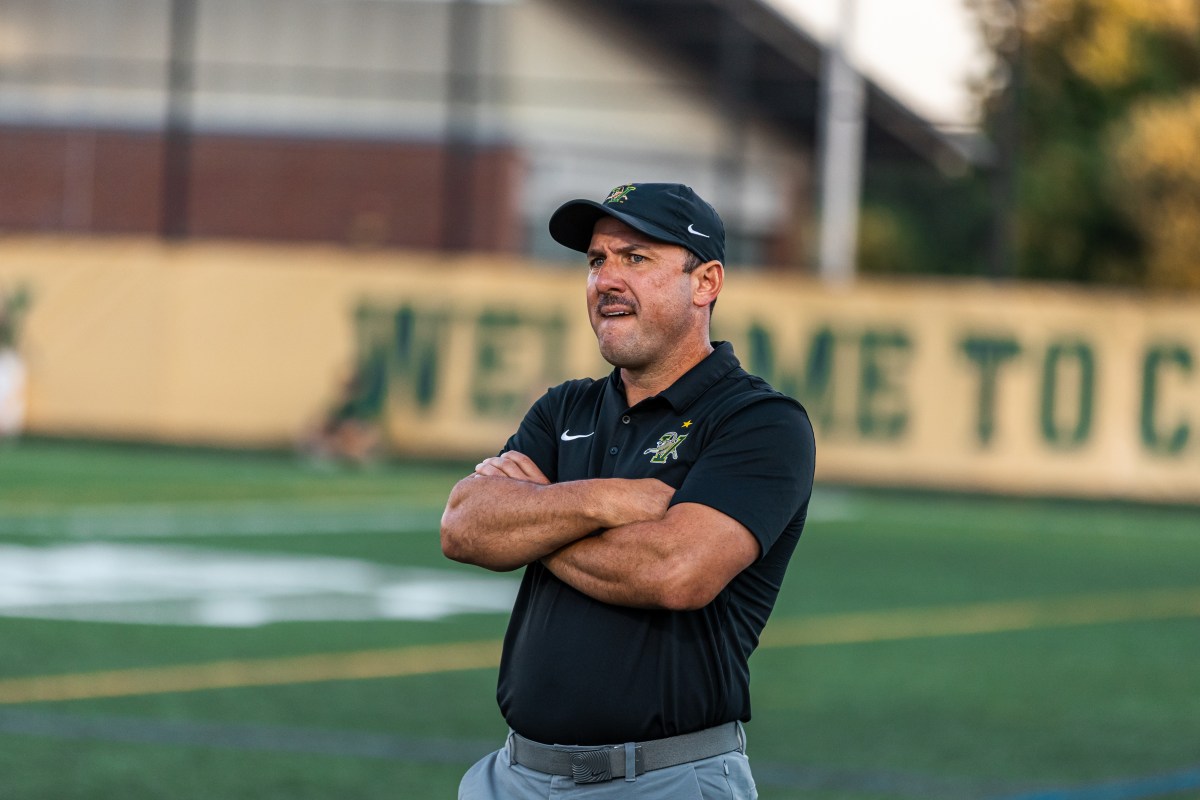 A coach wearing a black shirt and cap stands on a sports field with his arms crossed, looking ahead.