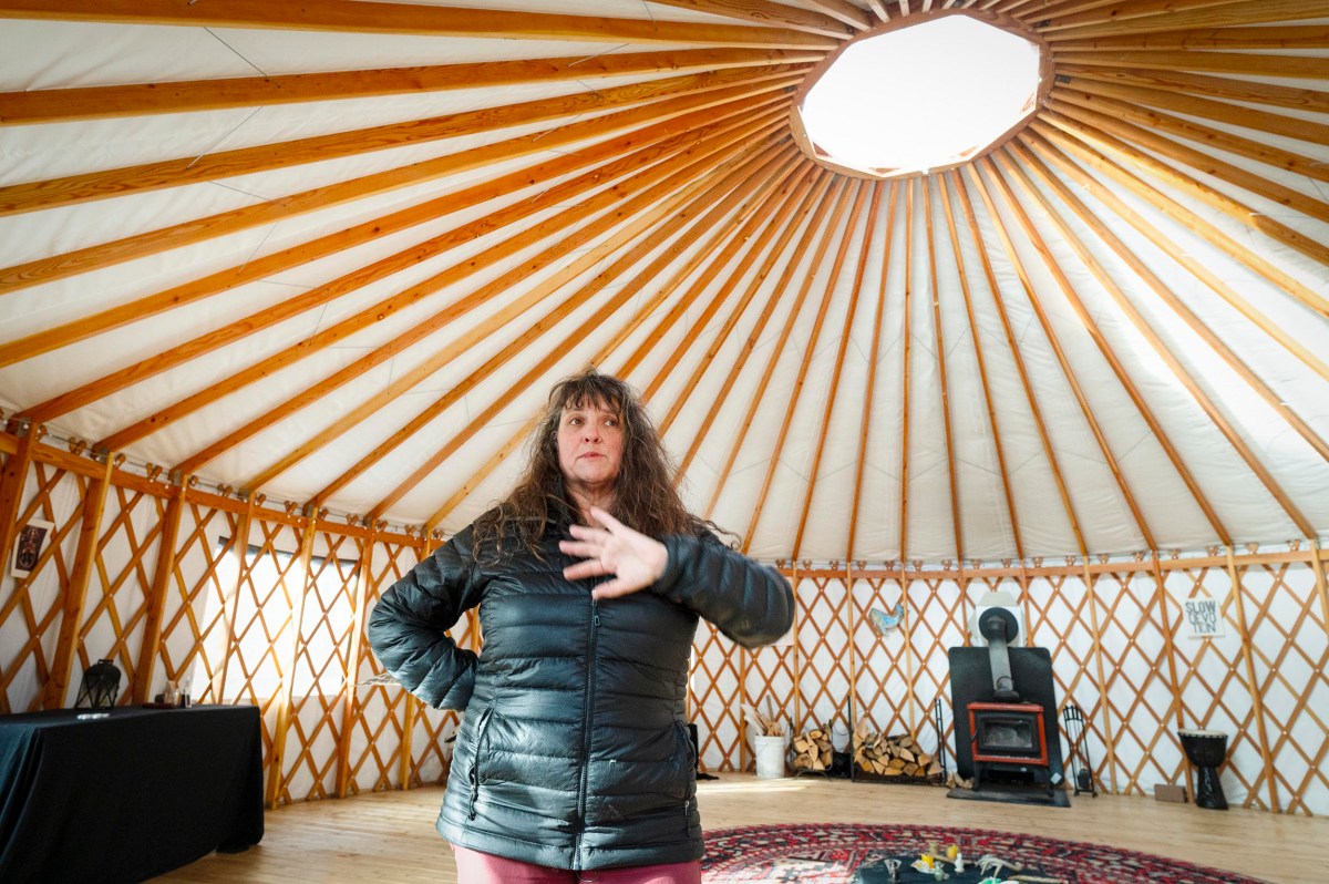 A woman in a black jacket stands inside a yurt with wooden beams, a circular skylight, and a wood stove in the background.
