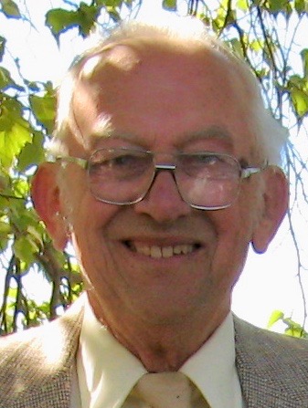 An older man with white hair and glasses smiles outdoors, wearing a light-colored shirt, tie, and jacket. Green leaves and branches are visible in the background.